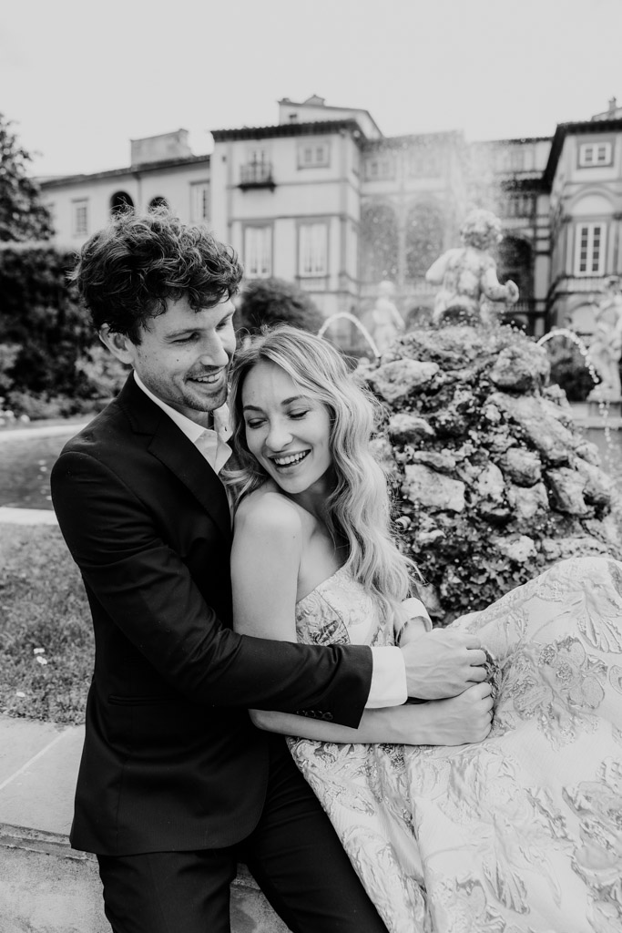 Black and white photo of couple in formal attire embracing and laughing by a fountain in a garden in Lucca, Italy