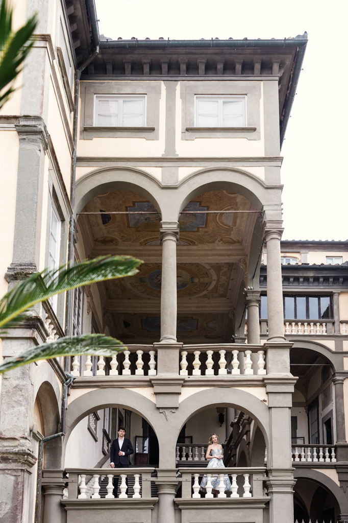 A couple stands apart on a grand, arched stone balcony of a historic building with painted ceilings in Lucca, Italy