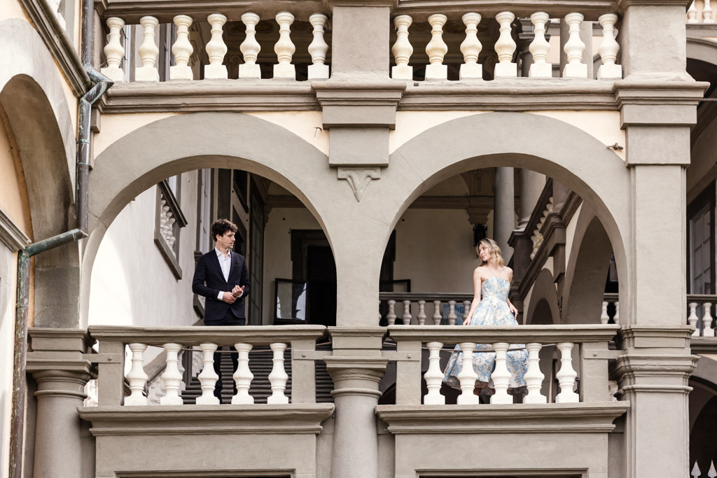 A man and woman stand apart on a balcony with arched openings in an elegant, historic building, during their Lucca engagement session