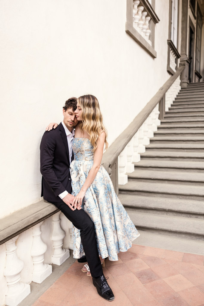 Couple poses by a staircase in Lucca, Italy; she wears a blue floral dress, he wears a dark suit