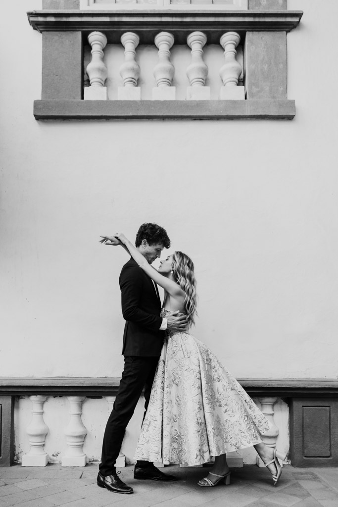 Black and white photo of couple danicng close in elegant attire in Lucca, Italy against a white wall adorned with decorative railings