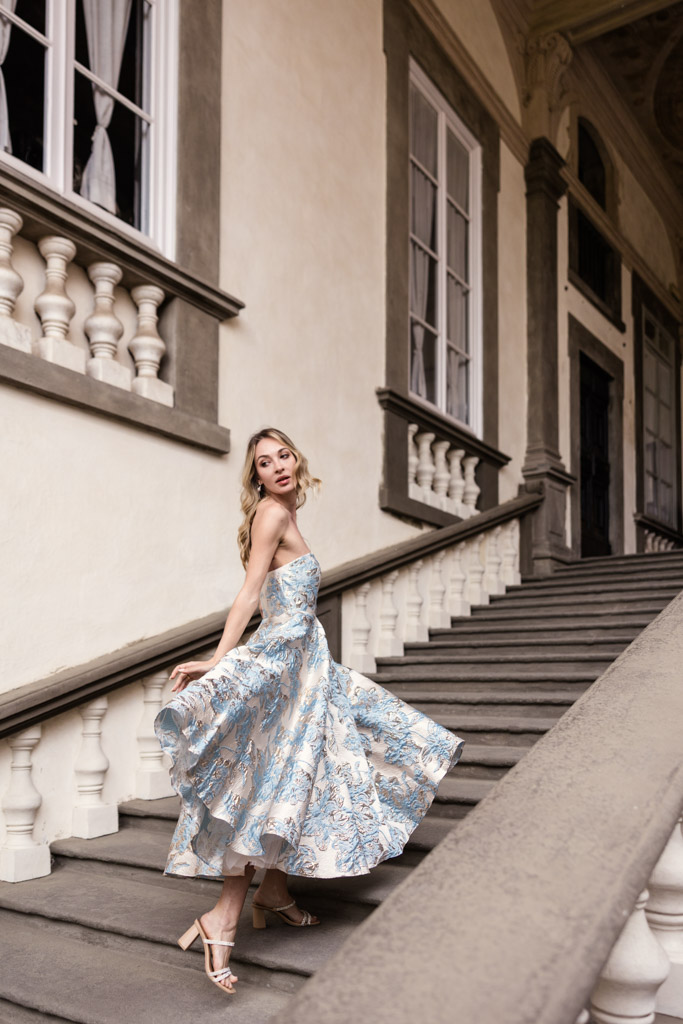 A woman in a flowing, patterned dress poses gracefully on elegant stone stairs by tall windows, in Lucca, Italy