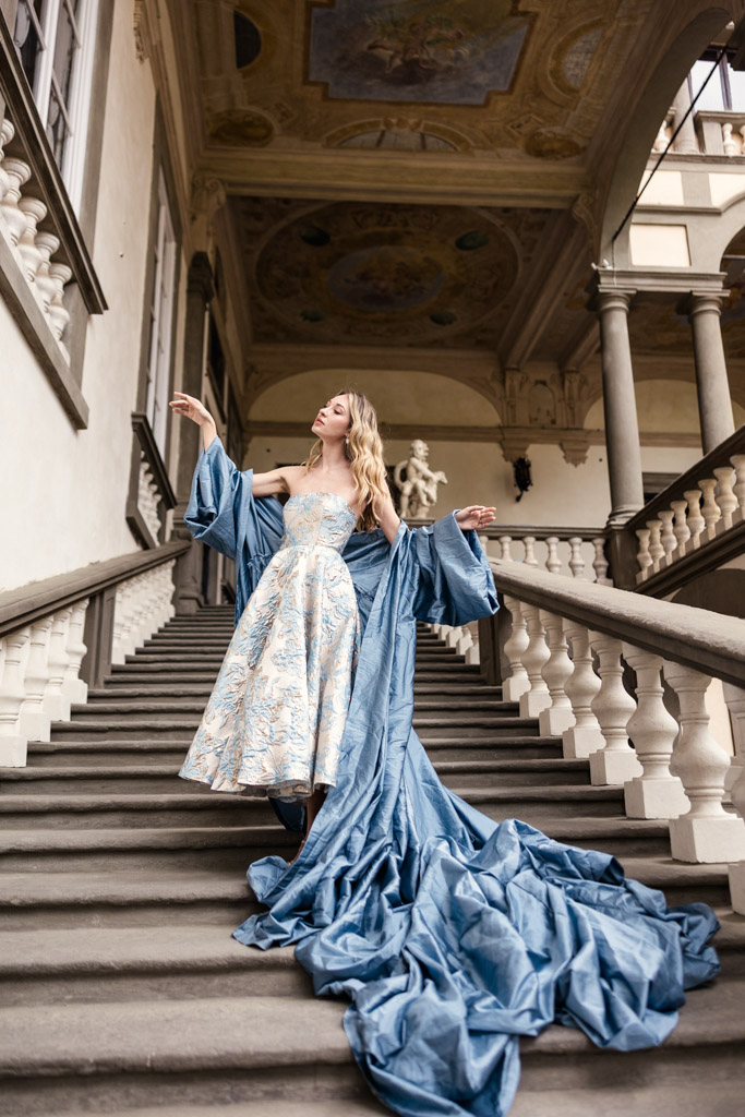 Woman in a flowing blue gown poses dramatically on a grand staircase in an ornate, historic building in Lucca, Italy