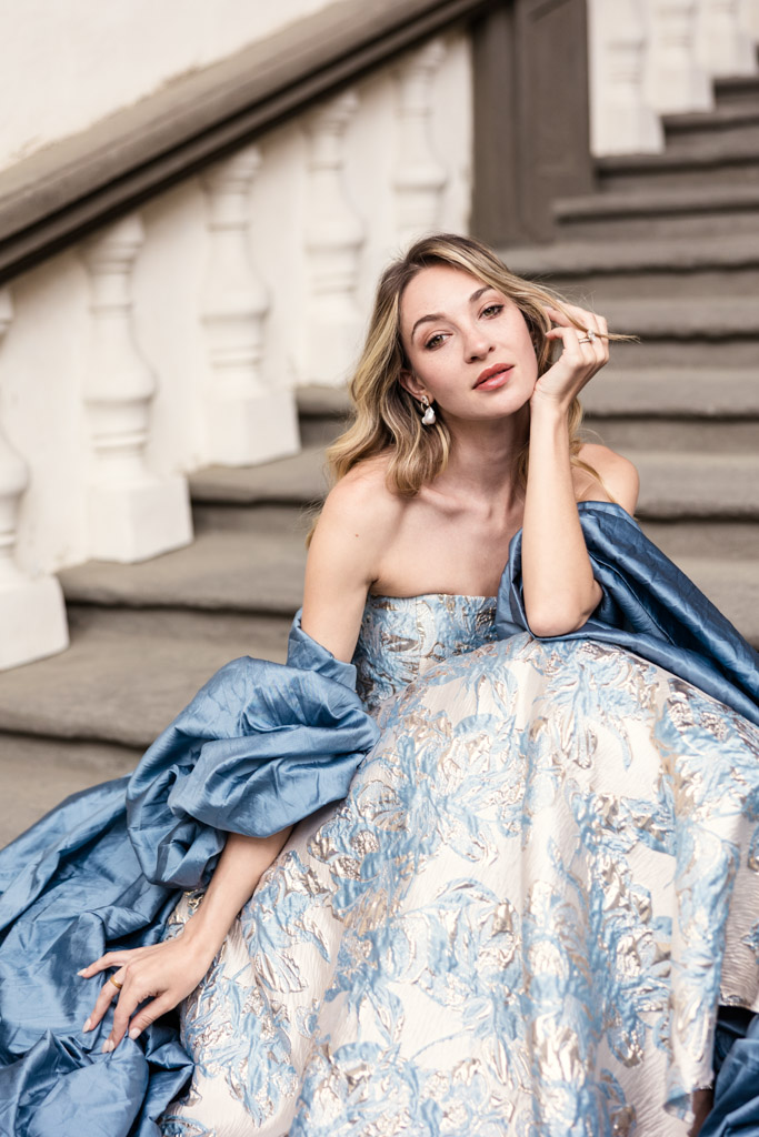 Portrait of woman in an elegant blue and silver gown on stairs in Lucca, Italy, resting her head on her hand and looking at the camera