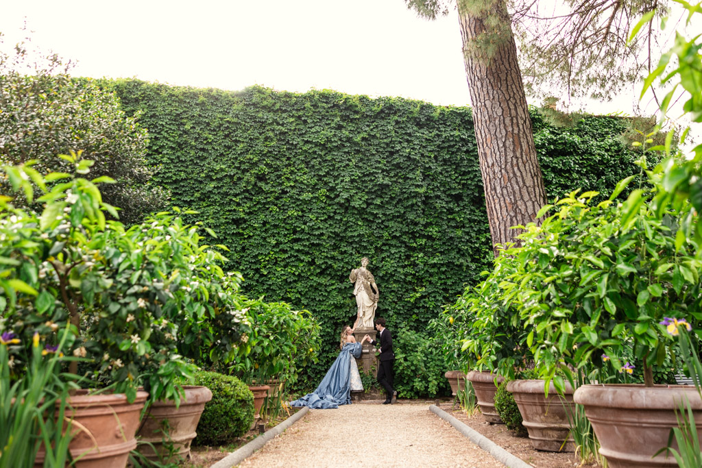 A couple stands by a statue in Orto Botanico Comunale di Lucca, surrounded by greenery and large potted plants