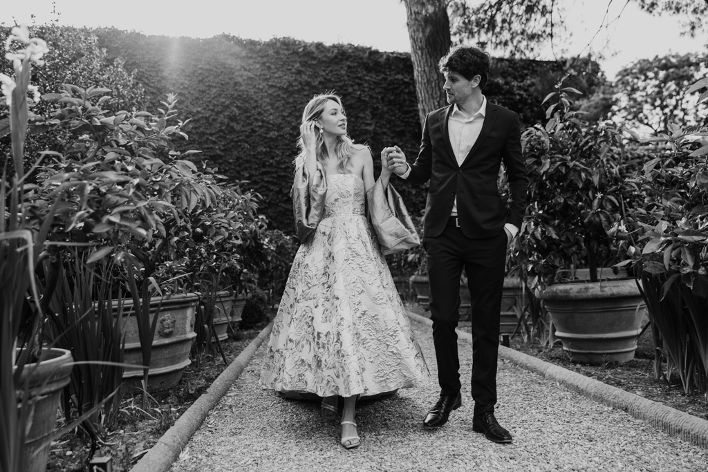 Black and white photo of couple dressed formally walking hand in hand on a garden path in Orto Botanico Comunale di Lucca surrounded by potted plants