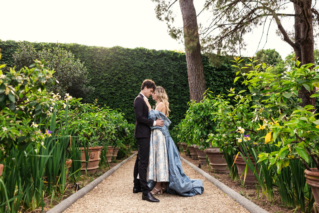 A couple dressed formally embraces on a path in Orto Botanico Comunale di Lucca surrounded by lush greenery and potted plants