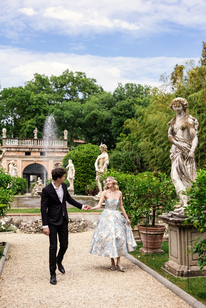 A couple walks hand in hand through Orto Botanico Comunale di Lucca with statues and greenery under a partly cloudy sky