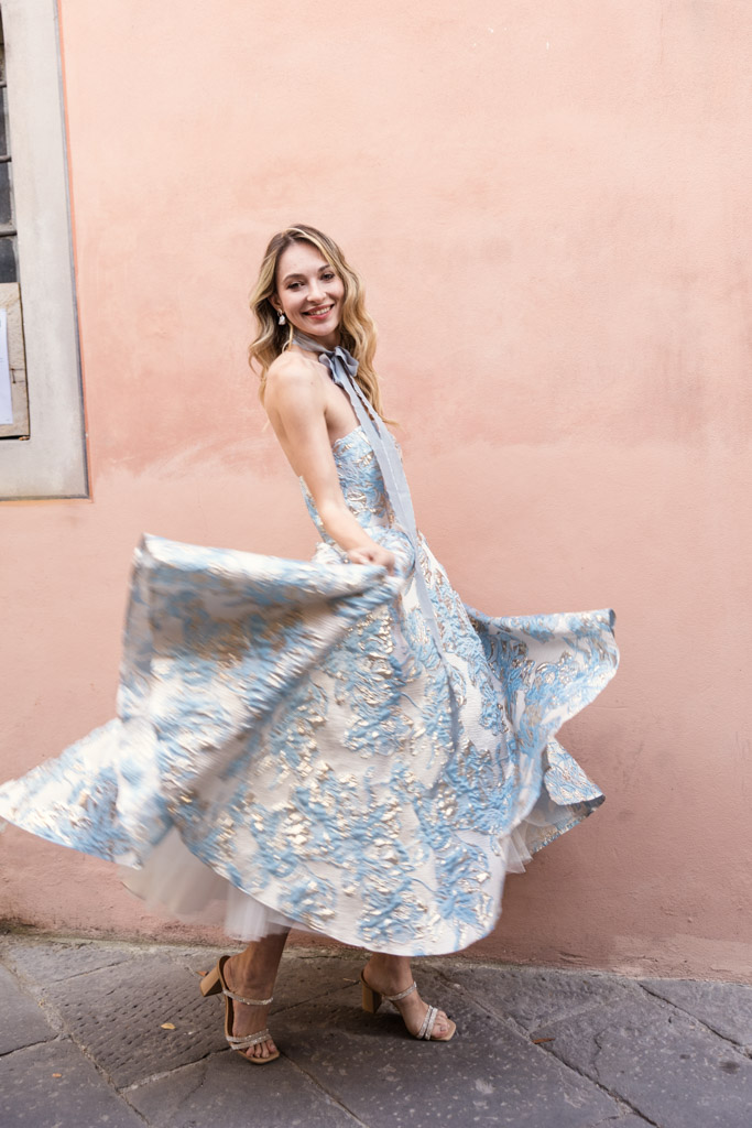 A woman in a flowing blue and silver dress twirls, smiling, against a peach-colored wall in Lucca, Italy