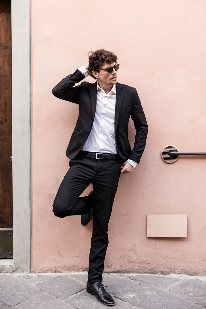 Man in a black suit and sunglasses leans against a pink wall in Lucca, Italy with one hand on his head and one leg raised