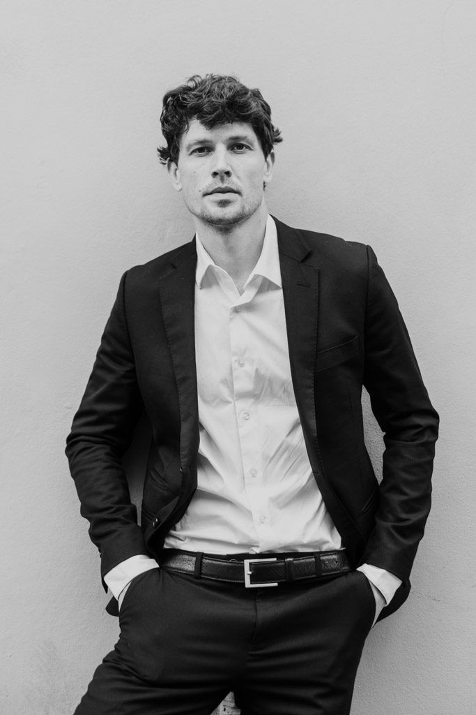 Black and white portrait of man in a suit and open-collared shirt against a plain wall, hands in pockets