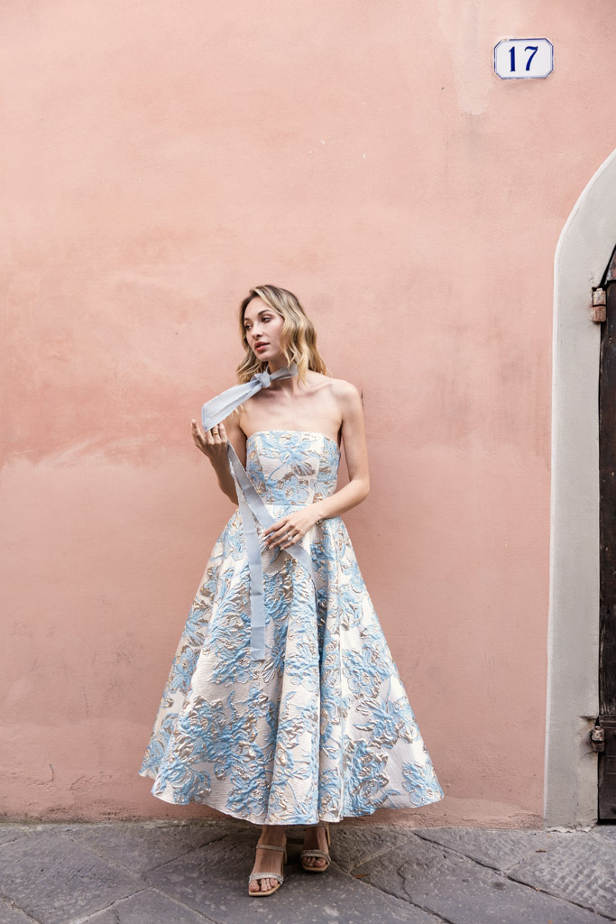 Woman in a strapless blue floral dress stands by a peach-colored wall under a street number 17 sign in Lucca, Italy