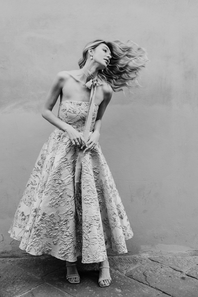 Black and white photo of woman in a strapless floral dress flipping her hair while standing against a plain wall