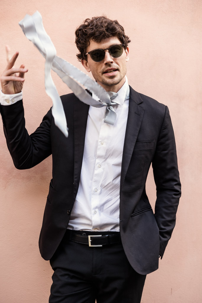 Man in a black suit and sunglasses tossing a silver tie, standing against a pink wall
