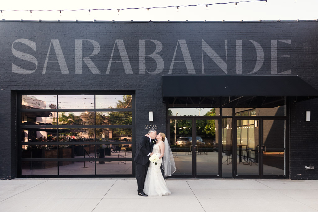 Bride and groom kiss outside Sarabande Chicago, framed by its iconic sign and modern glass garage doors during their summer wedding celebration