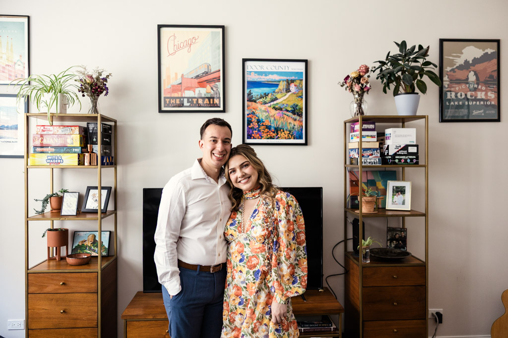 Portrait of happy couple together in their living room with shelves, plants, and framed art on the walls during their summer engagement session