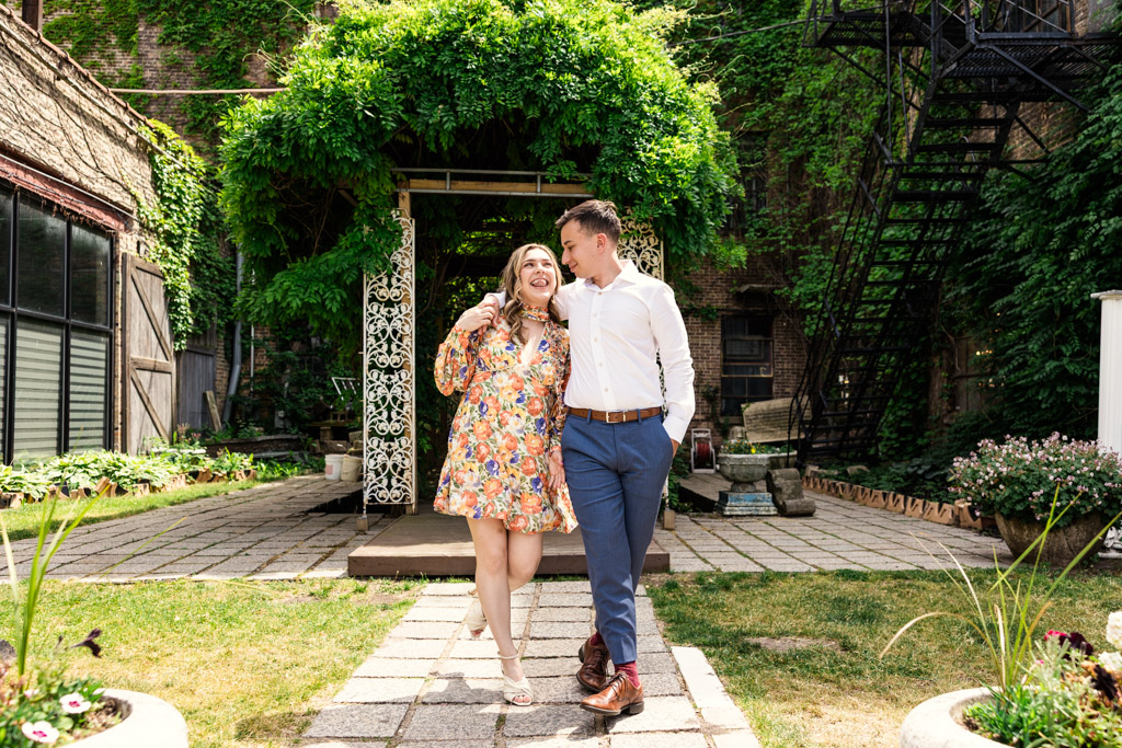 Happy engaged couple walks arm in arm on a garden path, surrounded by greenery with a gazebo in the background outside their Salvage One