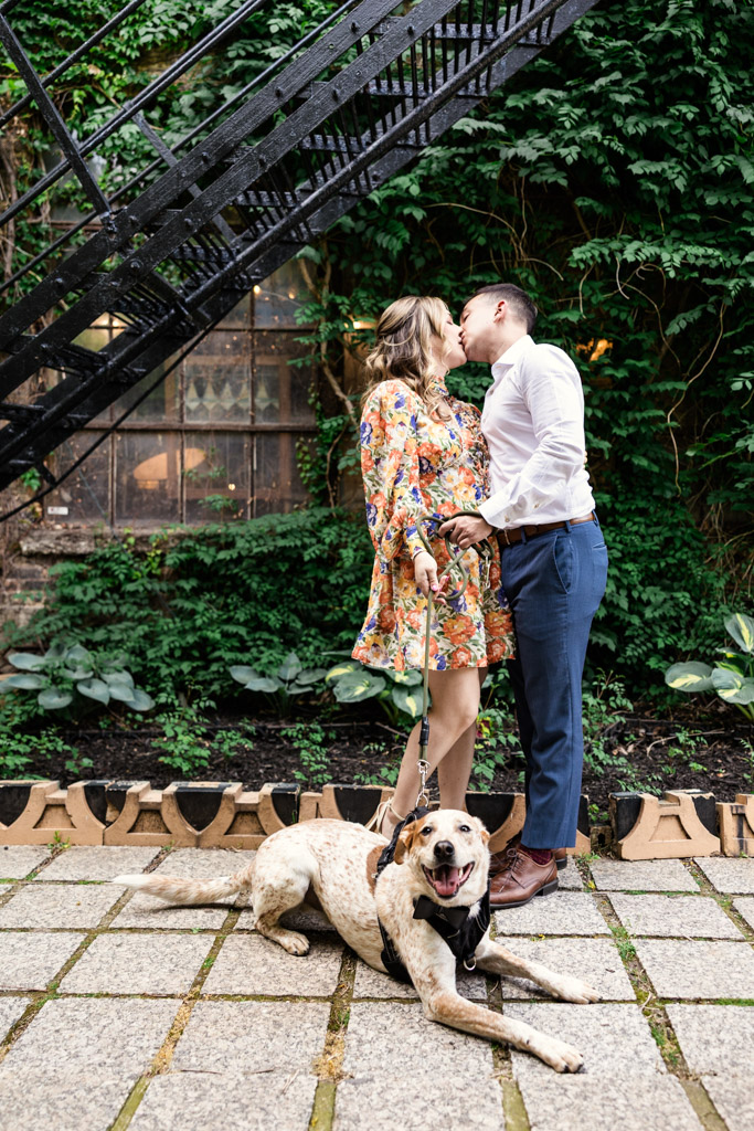 Engaged couple kisses outside Salvage One with their dog lying at their feet