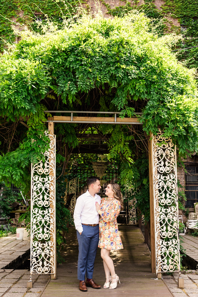 Engaged couple stands smiling while holding each other under a leafy archway outside Salvage One