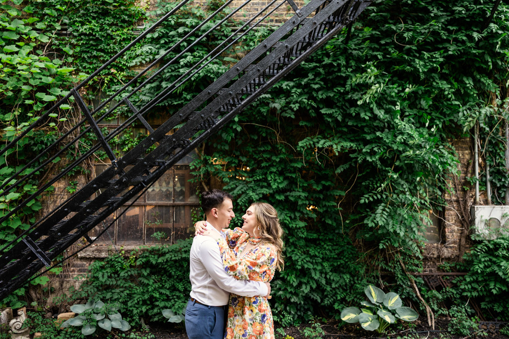 Engaged couple embraces under a metal staircase outside Salvage One, surrounded by lush green vines on a brick wall