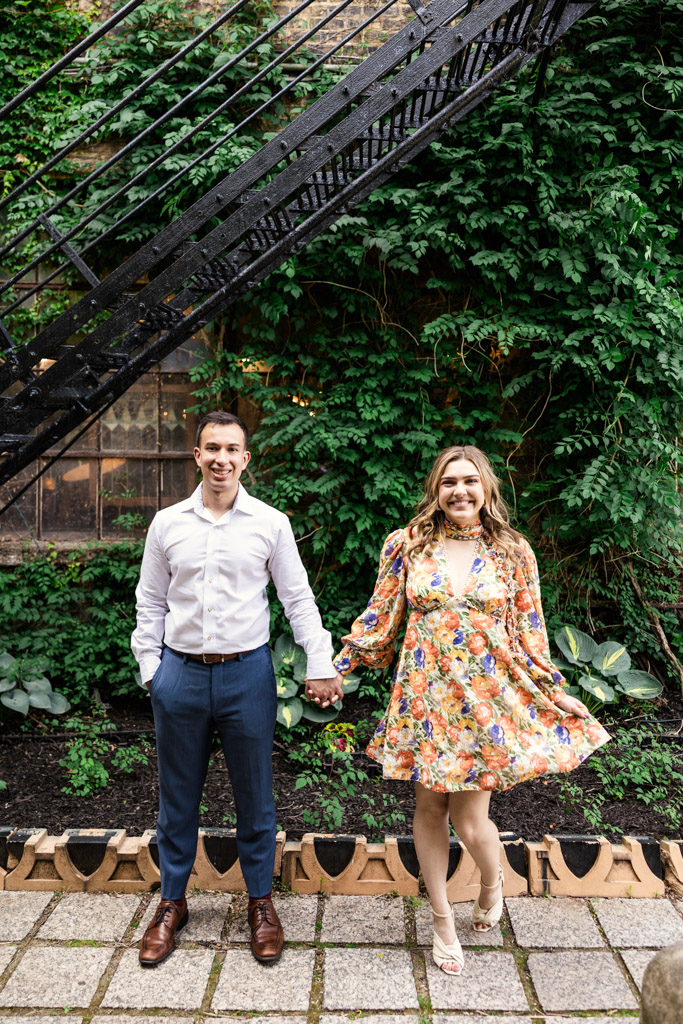 A couple holds hands and smiles, standing under a metal stairway by a green, leafy wall during their Salvage One engagement session