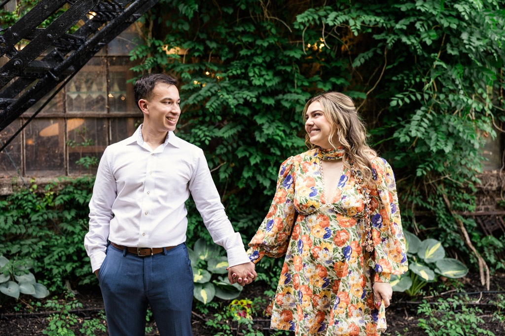 A smiling couple holding hands stands looking at each other warmly in front of an ivy-covered wall during their Salvage One engagement session