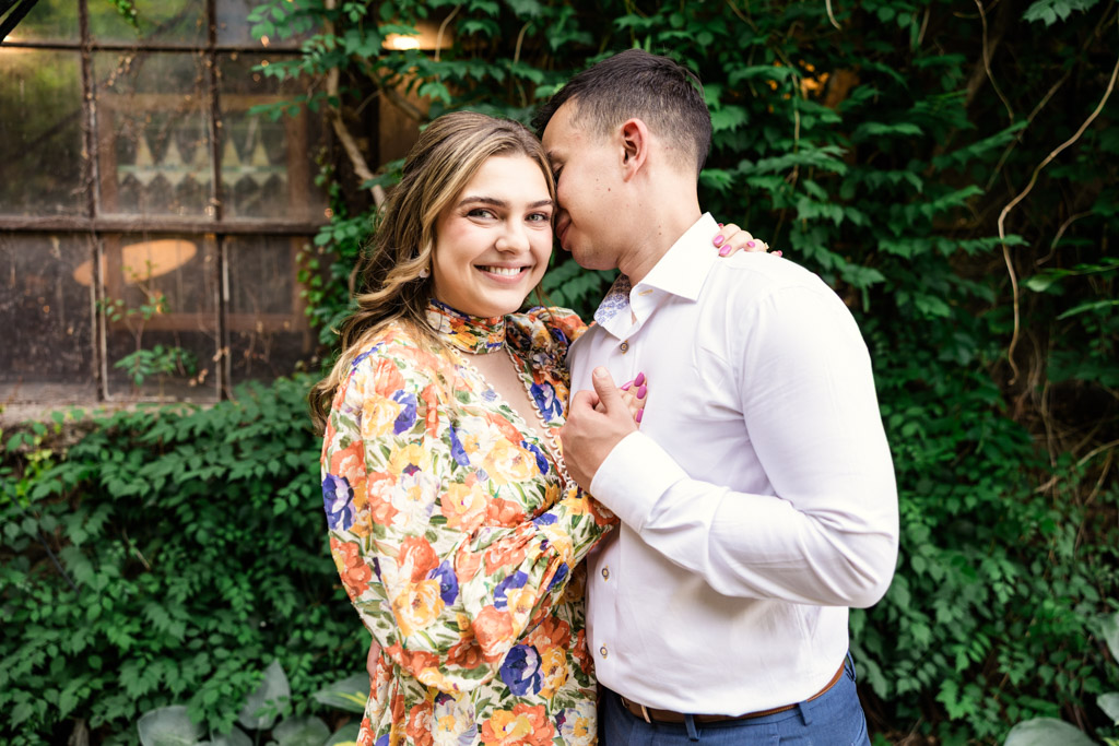A smiling woman in a floral dress hugs her partner in a white shirt, surrounded by lush greenery during their Salvage One engagement session