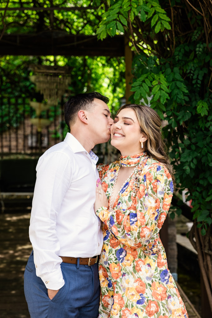 Man kisses his partner on the cheek as they stand outdoors under green leafy vines; she wears a floral dress during their Salvage One engagement session
