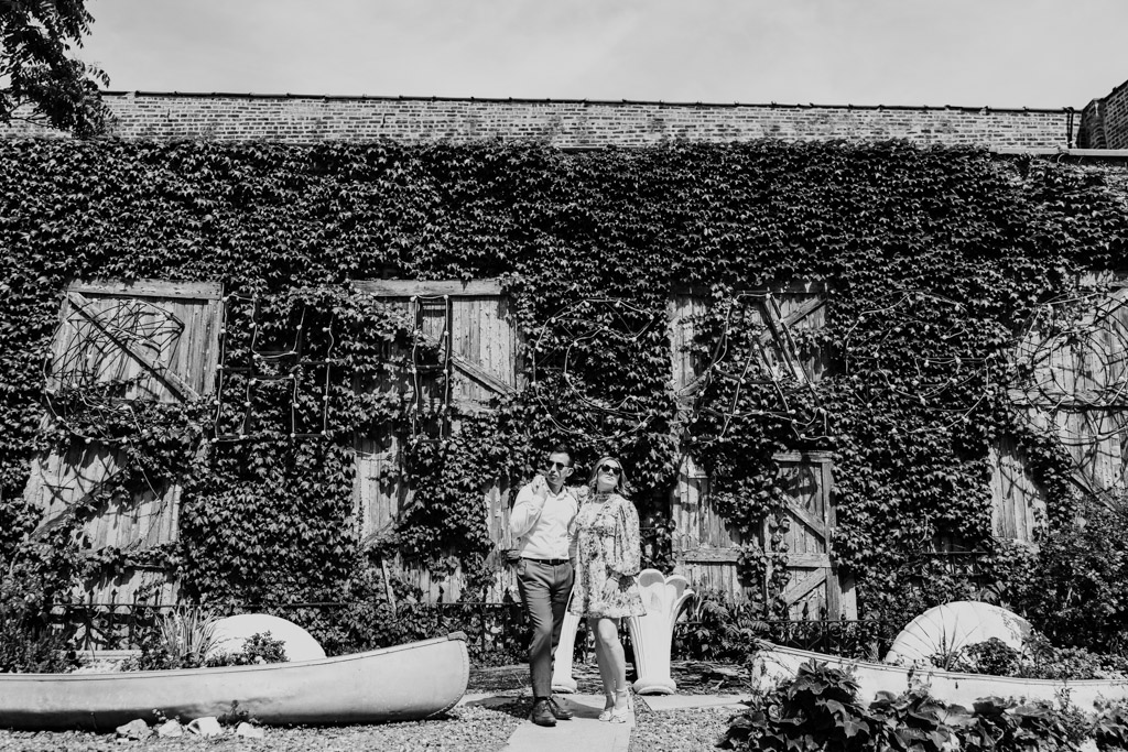 Black and white photo of engaged couple posing together in front of a vine-covered wall with vintage outdoor decor outside Salvage One