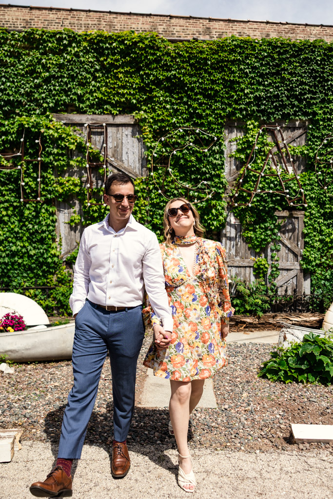 A couple in sunglasses holds hands and smiles while walking outdoors in front of a wall covered in green ivy outside Salvage One during their summer engagement session