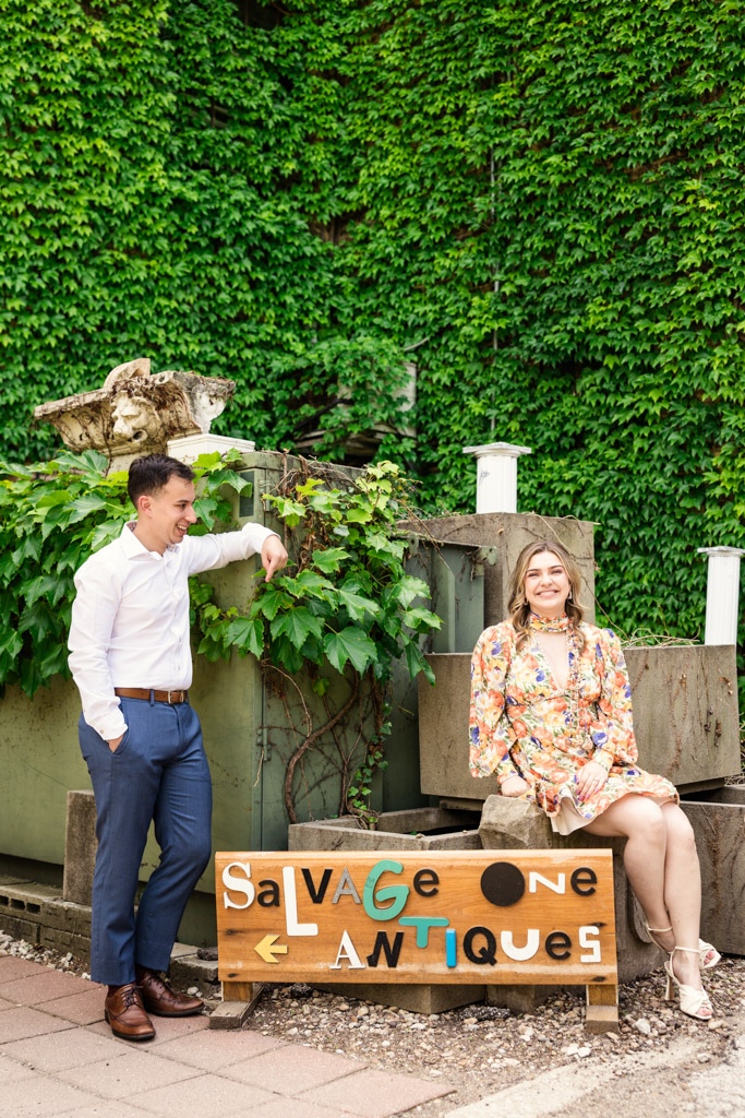 A man and woman pose by a colorful sign in front of leafy green ivy during their Salvage One engagement session