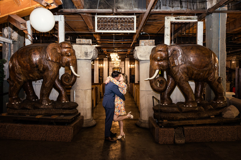 A couple kisses between two large wooden elephant statues in the warmly lit indoor space of Salvage One during their summer engagement session