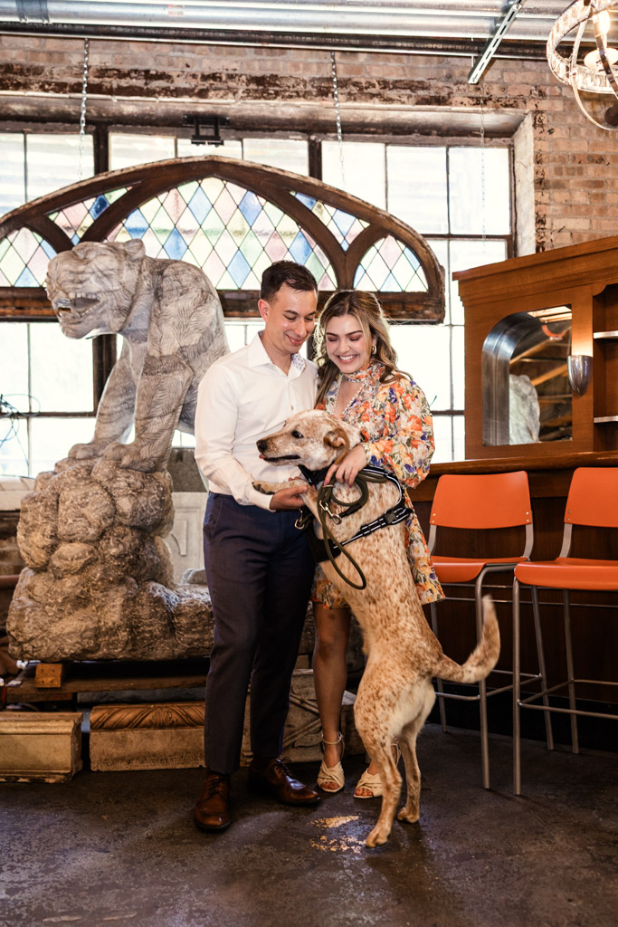 Happy couple smiles as their dog stands on its hind legs inside a rustic room with stained glass windows during their Salvage One engagement session