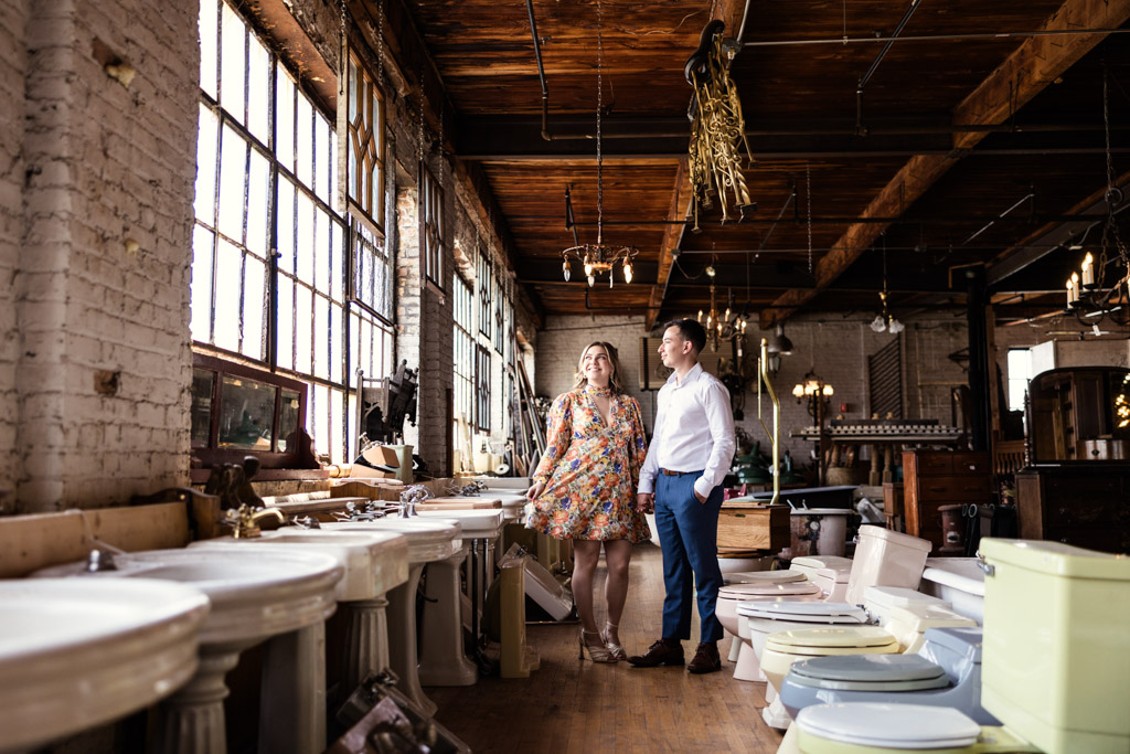 Engaged couple stands together in a vintage shop filled with sinks and fixtures, sunlight streaming through large windows at Salvage One