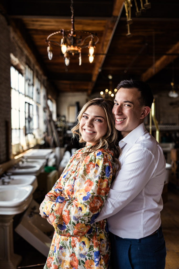 Portrait of happy engaged couple embracing in a rustic, sunlit room with wooden ceilings and antique fixtures at Salvage One