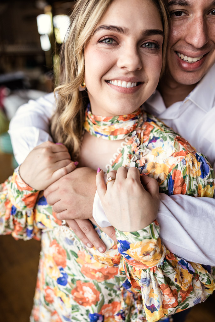 Happy engaged couple embraces during their Salvage One engagement session, with the woman displaying her engagement ring and wearing a colorful floral dress