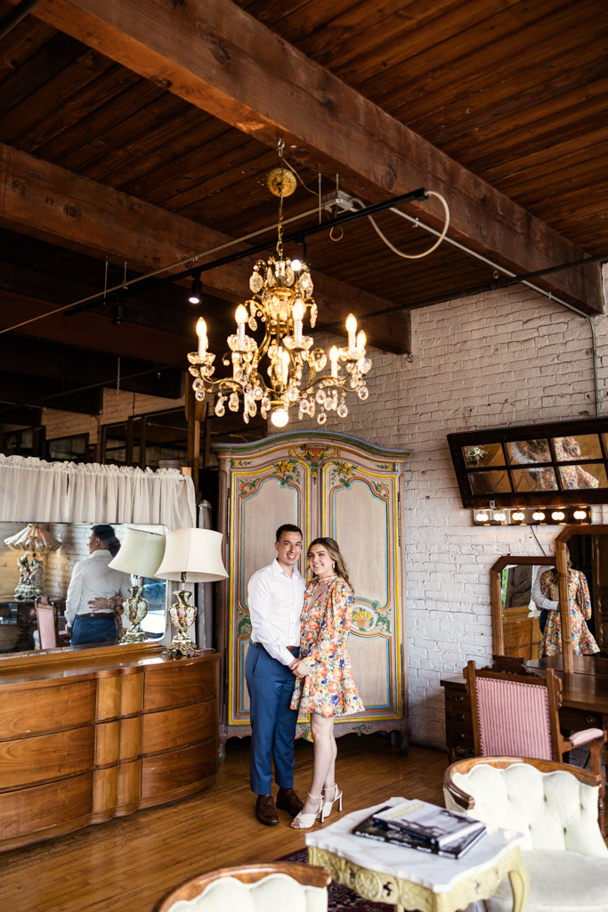 Engaged couple stands smiling in a vintage room at Salvage One, surrounded by wooden beams, chandeliers, and ornate furniture