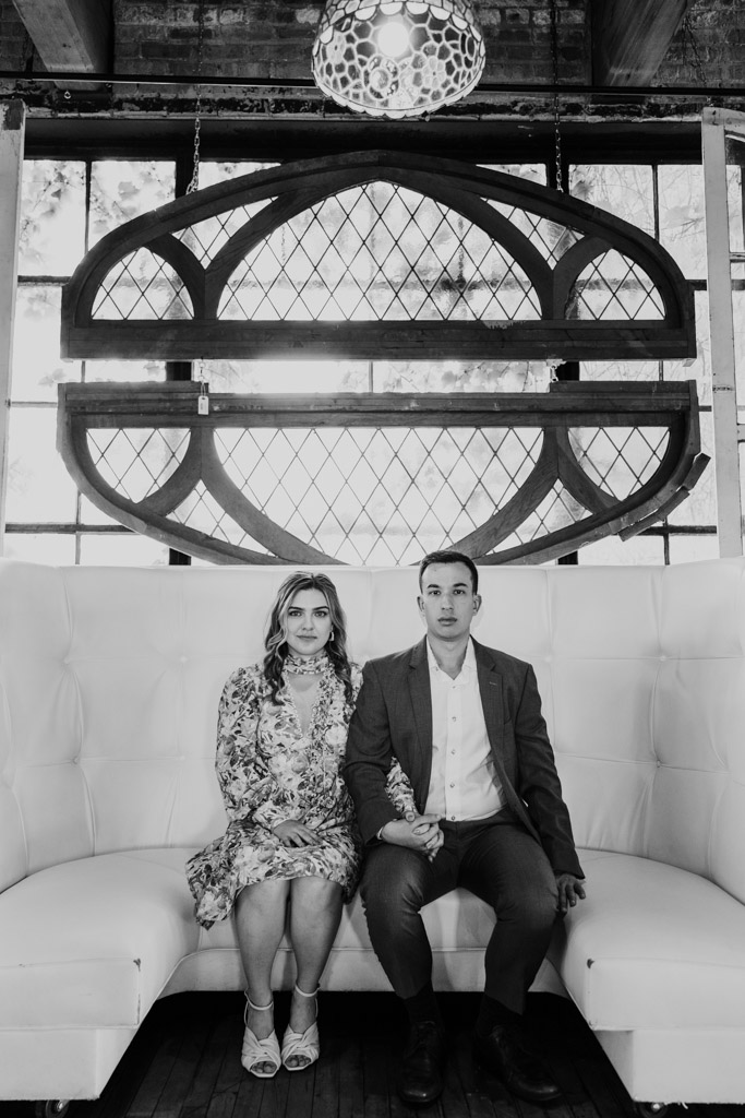 Black and white portrait of engaged couple sitting side by side on a white couch in front of decorative windows, looking at the camera during their Salvage One engagement session