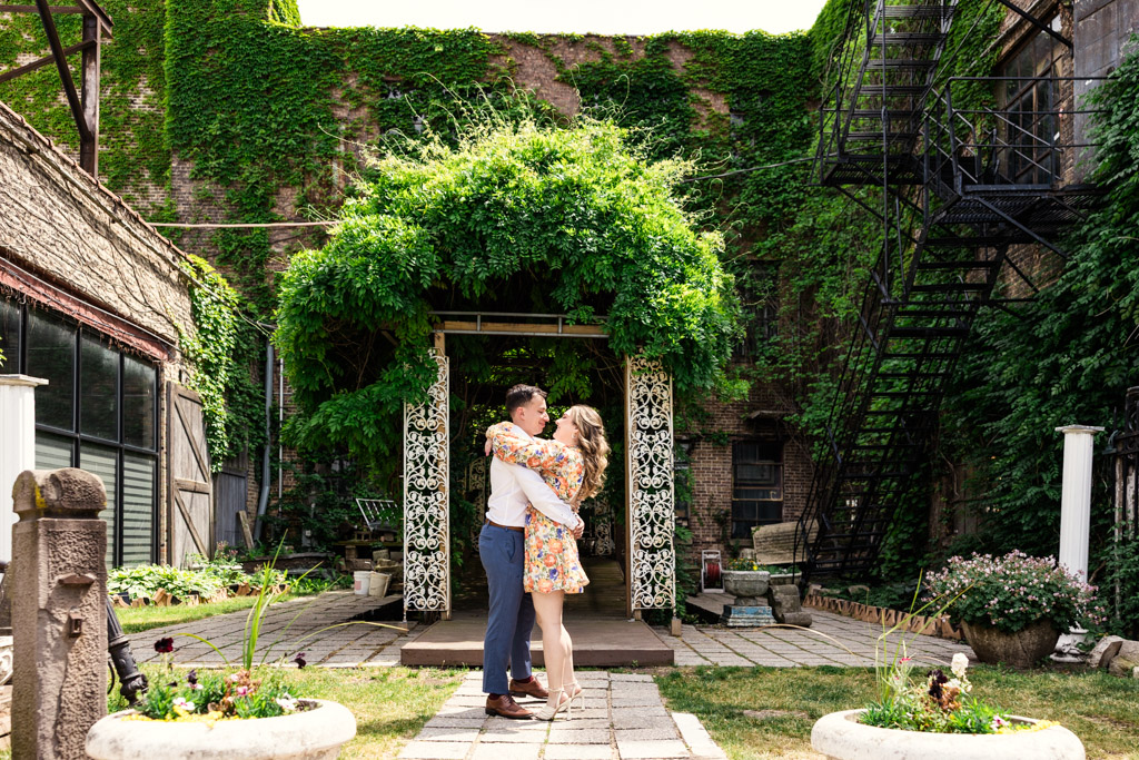 A couple embraces and smiles in a lush courtyard with ivy-covered walls and a decorative arbor outside Salvage One during their summer engagement session
