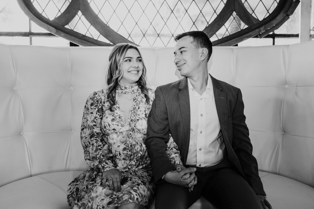 Black and white photo of happy engaged couple sitting on a white couch at Salvage One, holding hands and looking at each other affectionately