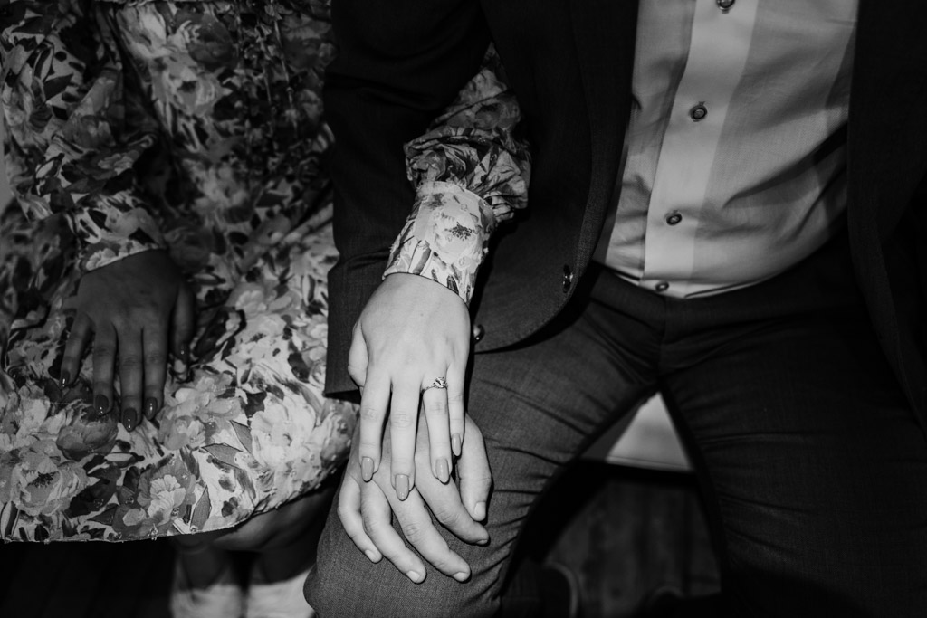 Black and white photo of engaged couple's hands as they sit close together inside Salvage One, the woman's hand showing off a sparkling ring
