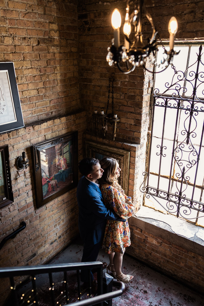 A couple stands by a window in a brick stairwell at Salvage One, embracing and looking outside through ornate iron bars, during their summer engagement session
