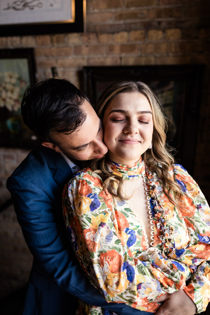 A man hugs a smiling woman from behind during their Salvage One engagement session; she wears a colorful floral dress and has her eyes closed