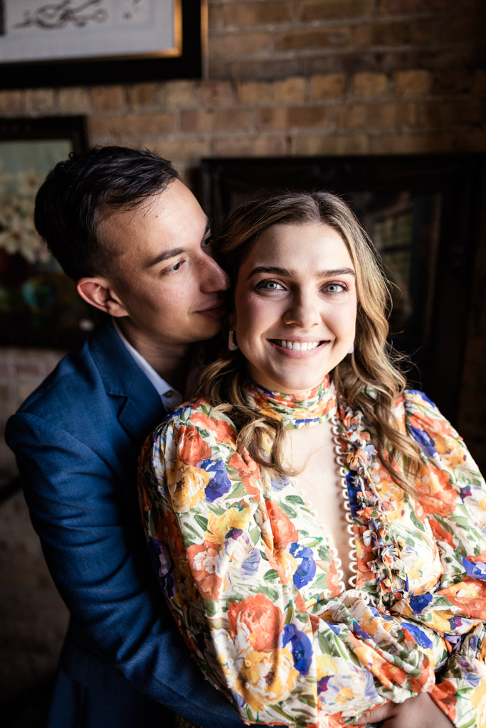 A smiling woman in a floral dress is hugged from behind by her partner in a blue suit, both indoors at Salvage One during their summer engagement session
