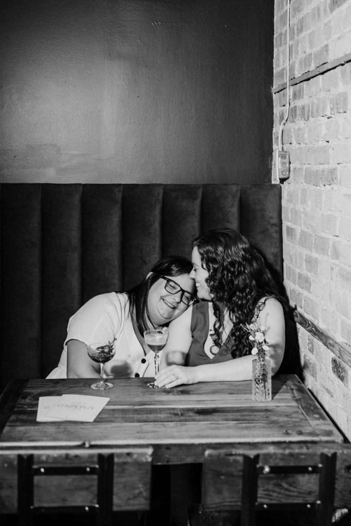Black and white photo of two women sitting close together at a wooden table inside Clara cocktail bar in Chicago during summer engagement session