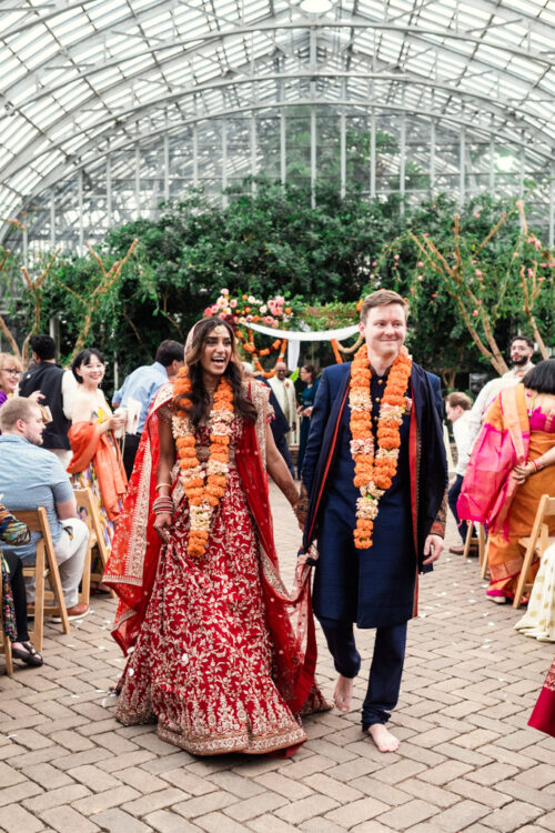 A bride and groom in traditional attire walk hand in hand at a vibrant indoor wedding ceremony.