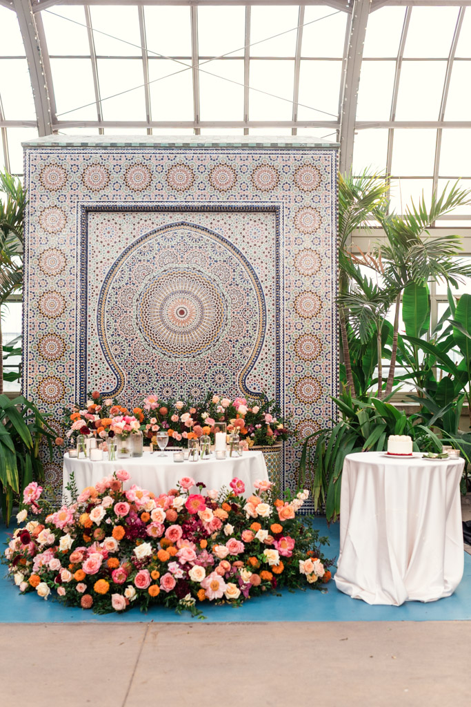 Sweetheart table with colorful floral arrangements and candles in front of a mosaic backdrop, with a small cake on a round table for Garfield Park Conservatory wedding reception