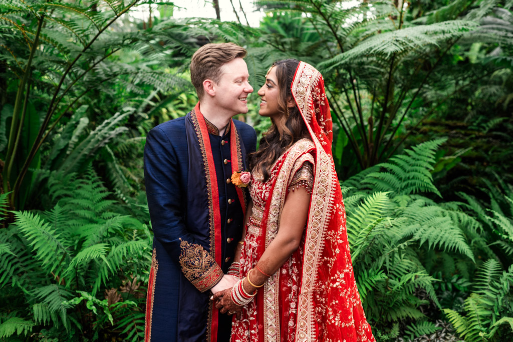 A couple in traditional Indian wedding attire stands together in front of lush green ferns, holding hands