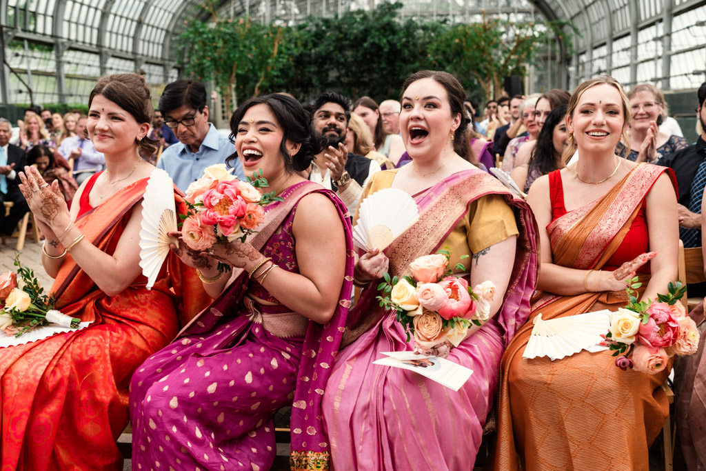 Four bridesaids in colorful saris laugh and clap while holding flowers at a Garfield Park Conservatory wedding ceremony