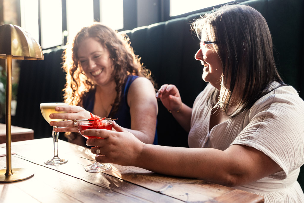 Two women sitting at a table in a sunlit Clara cocktail bar, smiling and holding cocktails during their Wicker Park engagement session
